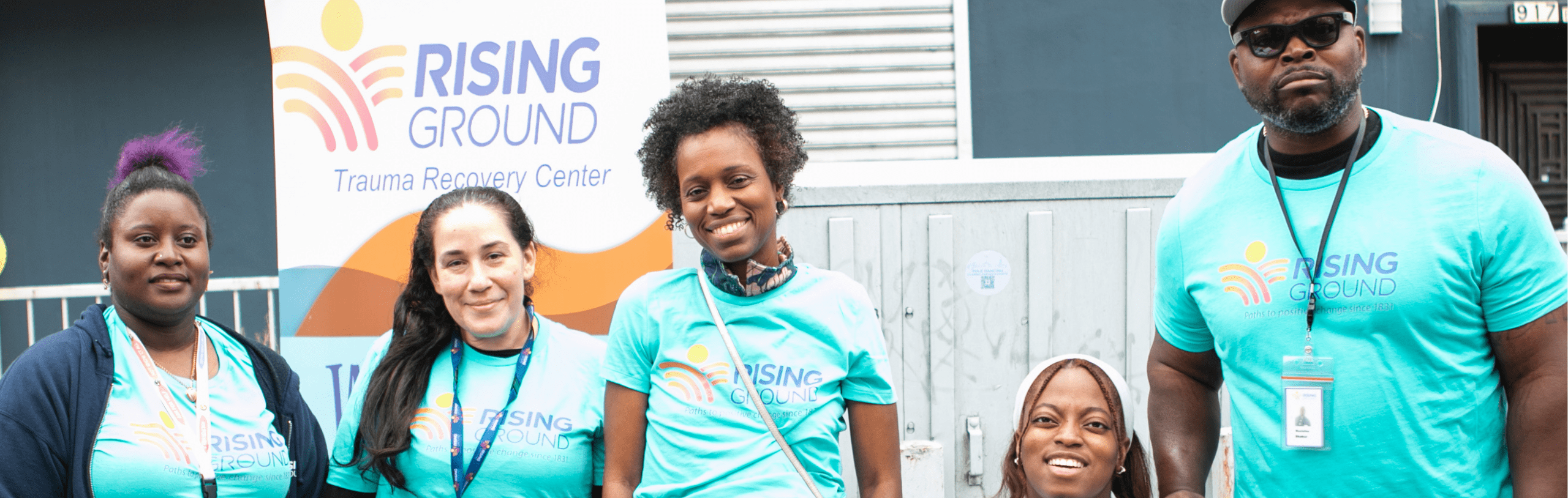 Five Rising Ground team members wearing blue shirts smile at the camera while standing outdoors in front of a Rising Ground sign.